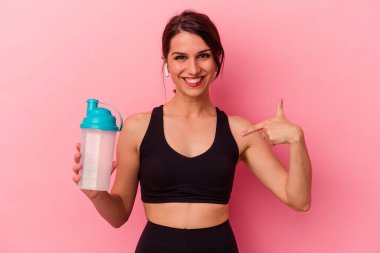 Pembe arka planda izole edilmiş protein içeceği içen beyaz bir kadın gömlek fotokopi alanını işaret ediyor, gururlu ve kendinden emin.