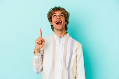 Mavi arka planda soyutlanmış beyaz makyajlı genç bir adam iki ön parmağı da boş bir alan gösteriyor..