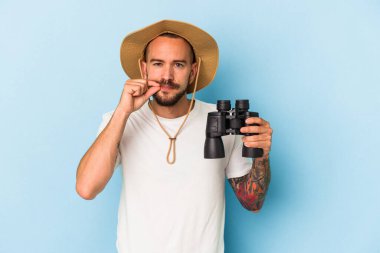 Dürbünü mavi arka planda tutan beyaz dövmeli genç bir adam. Parmakları dudaklarında bir sır saklıyor..