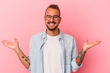 Pembe arka planda dövmeleri olan beyaz genç bir adam kollarıyla ölçek oluşturuyor, mutlu ve kendinden emin hissediyor..