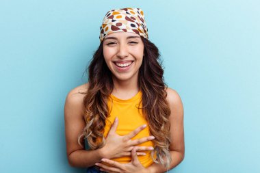 Mavi arka planda izole edilmiş genç Meksikalı kadın mutlu bir şekilde gülüyor ve ellerini karnının üstünde tutarak eğleniyor..