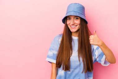 Pembe arka planda izole edilmiş genç beyaz kadın gülümsüyor ve başparmağını kaldırıyor.