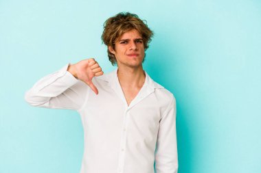 Makyajlı genç beyaz adam mavi arka planda izole edilmiş baş parmağını aşağı gösteren, hayal kırıklığı konsepti.