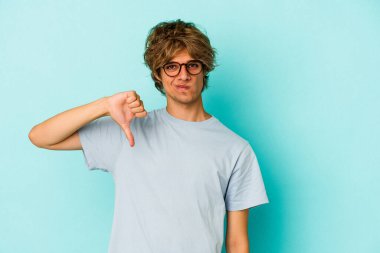 Makyajlı genç beyaz adam mavi arka planda izole edilmiş baş parmağını aşağı gösteren, hayal kırıklığı konsepti.