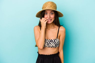 Genç, beyaz bir kadın kumsala gidiyor. Mavi arka planda izole edilmiş bir bikini giyiyor. Tırnaklarını yiyor..