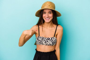 Mavi arka planda izole edilmiş bikini giyen beyaz bir kadın, gururlu ve kendinden emin bir şekilde, bir tişört fotokopi alanını işaret ediyor.