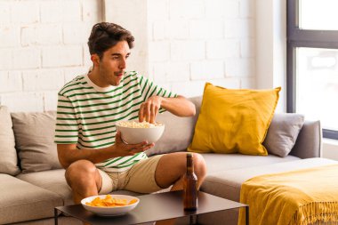 Genç melez adam kanepede oturmuş patlamış mısır yiyor.