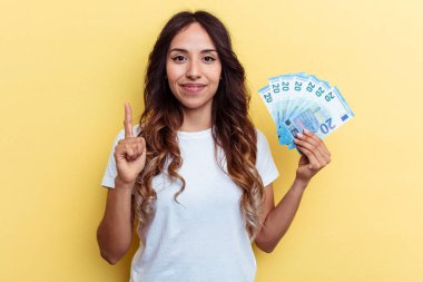 Sarı arka planda izole edilmiş banknotları tutan genç melez kadın parmak ile bir numarayı gösteriyor..