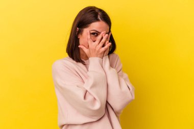 Sarı arka planda izole edilmiş genç beyaz kadın parmaklarının arasından korkmuş ve gergin bir şekilde göz kırp..