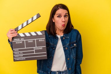 Sarı arka planda izole edilmiş bir el çırpma tahtası tutan genç beyaz kadın omuzlarını silker ve gözleri açık kalır..