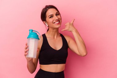 Pembe arka planda izole edilmiş protein içeceği içen genç beyaz kadın parmaklarıyla bir cep telefonu hareketi gösteriyor..