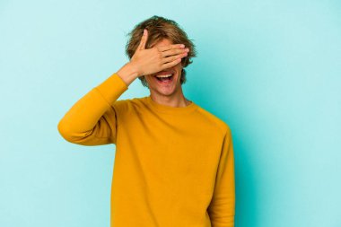 Mavi arka planda soyutlanmış beyaz makyajlı genç bir adam elleriyle gözlerini kapatıyor, gülümseyerek bir sürpriz bekliyor..