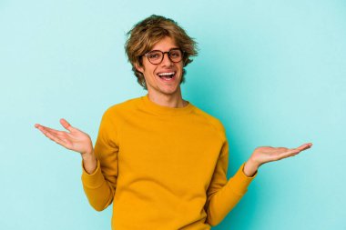 Mavi arka planda soyutlanmış beyaz makyajlı genç bir adam kollarıyla ölçeği yapıyor, mutlu ve kendinden emin hissediyor..