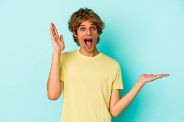 Mavi arka planda izole edilmiş beyaz makyajlı genç bir adam bir avuç içinde fotokopi alanı tutuyor, el üstünde tutuyor. Şaşırmış ve mutlu..