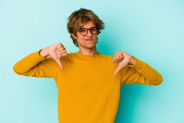 Makyajlı genç beyaz adam mavi arka planda izole edilmiş baş parmağını aşağı gösteren, hayal kırıklığı konsepti.