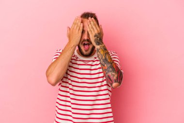Pembe arka planda dövmeleri olan beyaz bir adam yüzünün yarısını avuç içiyle örterek eğleniyor..