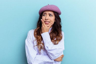 Mavi arka planda izole edilmiş genç Meksikalı kadın kafasının arkasına dokunuyor, düşünüyor ve bir seçim yapıyor..