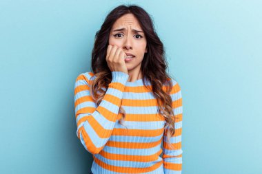 Mavi arka planda izole edilmiş genç Meksikalı kadın tırnaklarını yiyor, gergin ve çok endişeli..