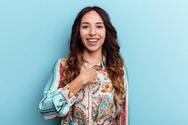 Mavi arka planda izole edilmiş genç Meksikalı kadın parmağını gösterip gülümseyerek şaşırdı..