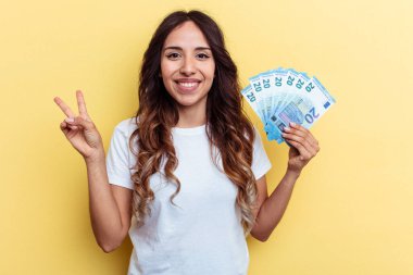 Sarı arka planda izole edilmiş banknotları tutan genç melez kadın neşeli ve kaygısız parmaklarıyla barış sembolü gösteriyor..