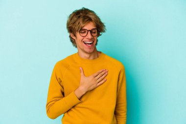 Mavi arka planda saklanmış beyaz makyajlı genç bir adam yüksek sesle gülüyor ve elini göğsüne koyuyor..