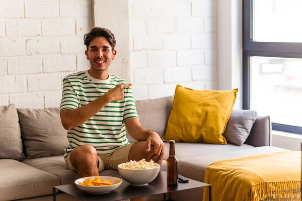 stock image Young mixed race man eating popcorns sitting on the sofa smiling and pointing aside, showing something at blank space.