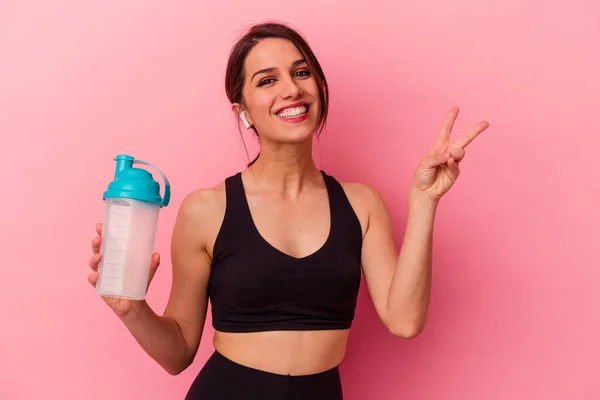 ピンクの背景に孤立したタンパク質シェイクを飲む若い白人女性喜びと屈託のない指で平和のシンボルを示す — ストック写真
