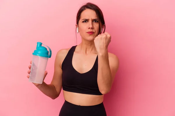 若い白人女性がピンクの背景に単離されたタンパク質シェイクを飲んでカメラに拳を示す 積極的な顔の表情 — ストック写真