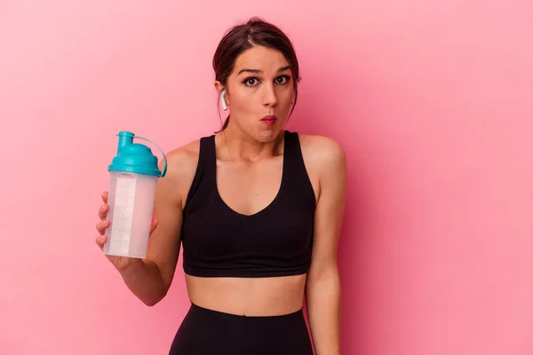 若いですCaucasian女性飲むタンパク質シェイク隔離ピンクの背景Shrugs肩とオープン目混乱 — ストック写真