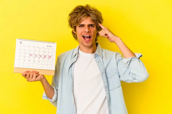 Young caucasian man with makeup holding calendar isolated on yellow background  showing a disappointment gesture with forefinger.