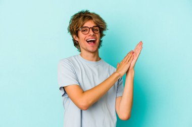 Makyajlı genç beyaz adam mavi arka planda izole edilmiş enerjik ve rahat hissediyor, ellerini ovuşturuyor kendinden emin..