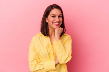 Pembe arka planda izole edilmiş genç beyaz kadın gülümsüyor ve kendinden emin, elleriyle çenesine dokunuyor..