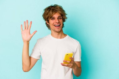 Makyajlı genç beyaz adam cep telefonunu mavi arka planda izole edip gülümseyerek beş numarayı parmaklarıyla gösterdi..