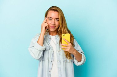 Elinde cep telefonu olan beyaz bir kadın mavi arka planda izole edilmiş. Şakağı işaret ediyor, düşünüyor ve bir göreve odaklanıyor..