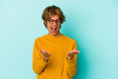 Makyajlı beyaz bir adam mavi arka planda izole edilmiş. Elinde avuç içi olan bir şey var ve kameraya sunuyor..