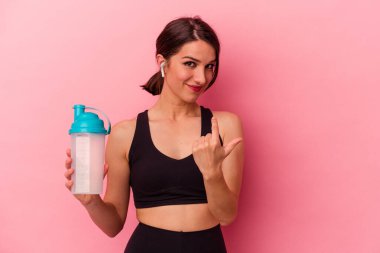 Pembe arka planda izole edilmiş protein içeceği içen genç beyaz kadın sanki seni davet ediyormuş gibi seni işaret ediyor..