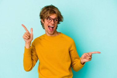 Makyajlı beyaz bir adam mavi arka planda izole edilmiş, farklı kopya alanlarına bakıyor, birini seçiyor, parmaklarıyla gösteriyor..