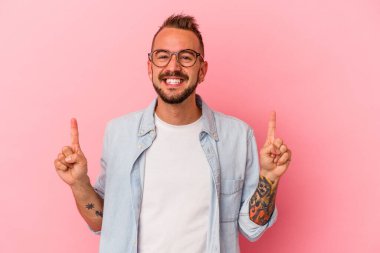 Pembe arka planda dövmeleri olan beyaz genç bir adam iki ön parmağı da boş bir alan gösteriyor..