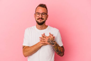 Pembe arka planda izole dövmeleri olan beyaz bir genç, dost bir ifadeye sahip, avuçlarını göğsüne bastırıyor. Aşk konsepti.