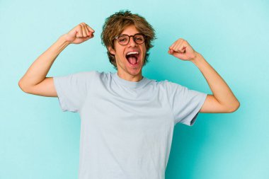 Mavi arka planda soyutlanmış beyaz makyajlı genç bir adam kollarıyla güçlü bir hareket gösteriyor, kadınsı gücün sembolü.