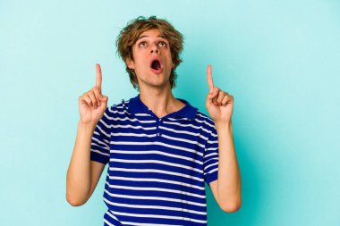 Makyajlı genç beyaz adam mavi arka planda izole edilmiş ağzı açık, üst tarafı işaret ediyor..