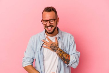 Pembe arka planda dövmeleri olan beyaz bir genç adam mutlu bir şekilde gülüyor ve ellerini karnının üstünde tutarak eğleniyor..