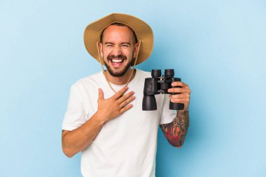 Mavi arka planda, elinde dürbün tutan beyaz dövmeli genç adam kahkahalarla gülüyor ve elini göğsünde tutuyor..