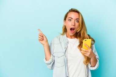 Elinde cep telefonu olan beyaz bir kadın mavi arka planda izole edilmiş ve yan tarafı işaret ediyor.