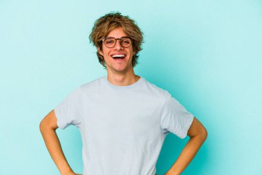 Makyajlı genç beyaz adam mavi arka planda izole edilmiş. Kendine güveni tam. Kalçalarını tutuyor..