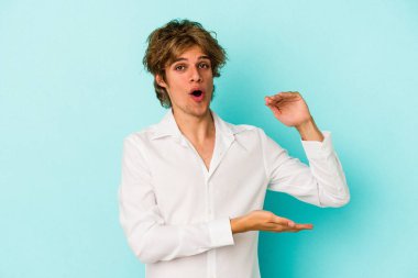 Mavi arka planda soyutlanmış beyaz makyajlı genç bir adam iki eliyle bir şey tutuyor..