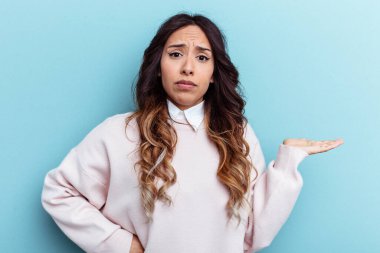 Meksikalı genç bir kadın mavi arka planda izole edilmiş. Sorgulama sırasında omuz silkiyor ve şüphe ediyor..