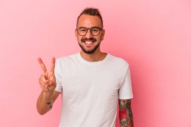 Pembe arka planda dövmeleri olan genç beyaz adam zafer işareti gösteriyor ve gülümsüyor..