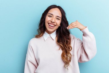 Mavi arka planda izole edilmiş genç Meksikalı bir kadın bir şeye gülüyor, elleriyle ağzını kapatıyor..