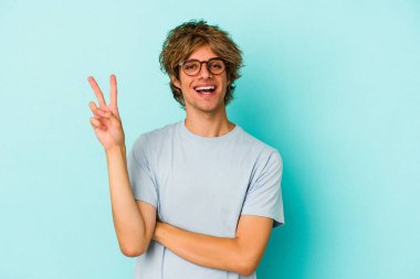 Makyajlı genç beyaz adam mavi arka planda iki numarayı parmaklarıyla gösteriyor..